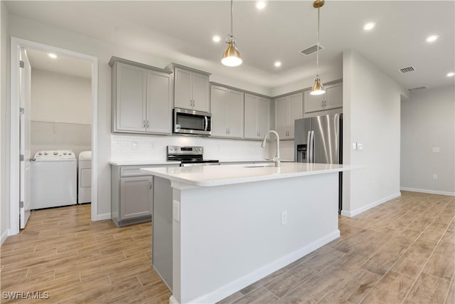 kitchen featuring washing machine and dryer, stainless steel appliances, gray cabinetry, and a center island with sink