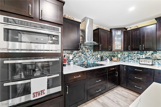 kitchen featuring black electric stovetop, wall chimney exhaust hood, stainless steel double oven, dark brown cabinetry, and light hardwood / wood-style floors