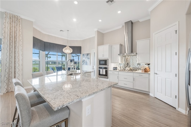 kitchen featuring a kitchen island with sink, white cabinets, wall chimney range hood, appliances with stainless steel finishes, and light hardwood / wood-style floors