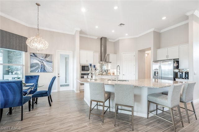 kitchen featuring stainless steel appliances, wall chimney range hood, sink, a large island with sink, and white cabinets