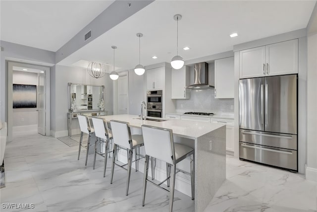 kitchen featuring wall chimney range hood, decorative light fixtures, a center island with sink, white cabinets, and appliances with stainless steel finishes