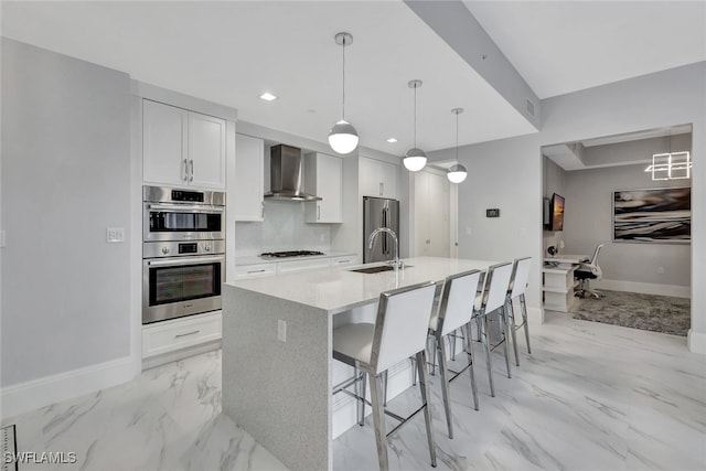 kitchen featuring wall chimney exhaust hood, pendant lighting, a kitchen island with sink, white cabinets, and appliances with stainless steel finishes