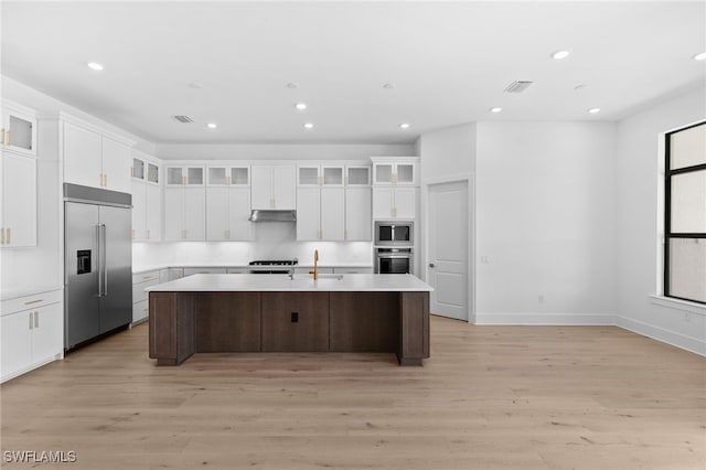 kitchen with light wood-type flooring, built in appliances, white cabinets, and a kitchen island with sink