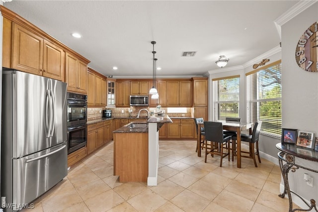 kitchen with dark stone countertops, crown molding, decorative light fixtures, a kitchen island with sink, and appliances with stainless steel finishes