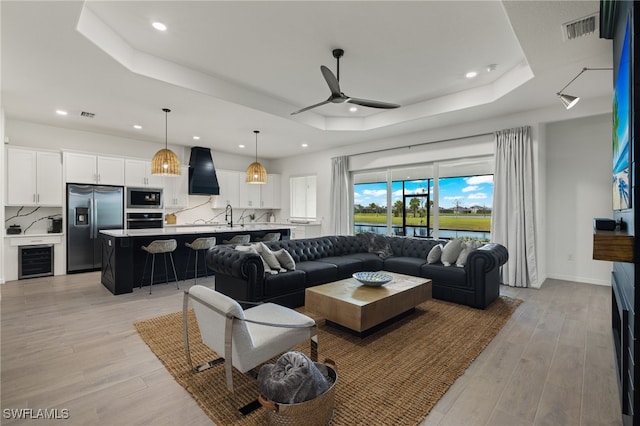 living room featuring a raised ceiling, ceiling fan, sink, light hardwood / wood-style floors, and wine cooler