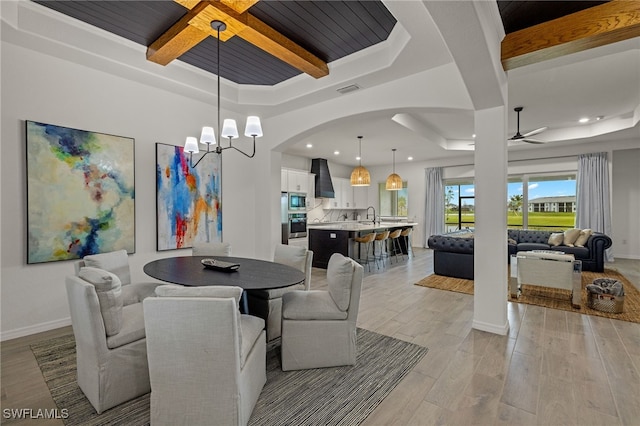 dining area with beamed ceiling, a raised ceiling, ceiling fan with notable chandelier, and light hardwood / wood-style flooring