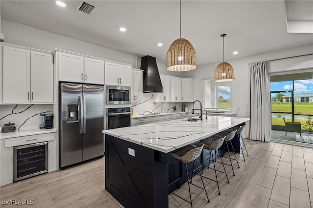 kitchen featuring premium range hood, an island with sink, appliances with stainless steel finishes, white cabinetry, and beverage cooler