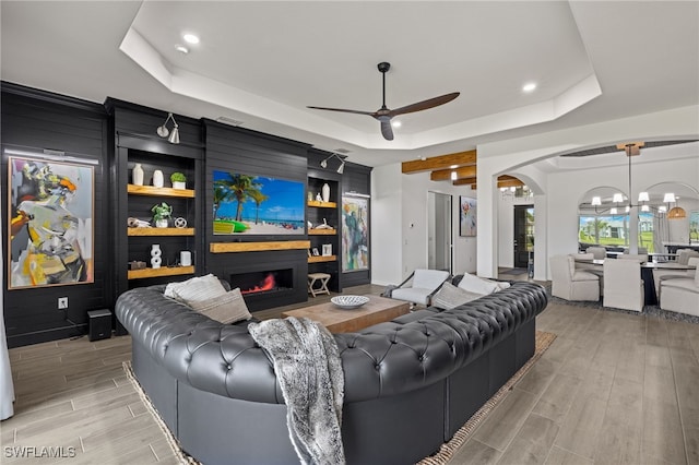 living room with ceiling fan with notable chandelier, wood walls, a raised ceiling, and light hardwood / wood-style flooring