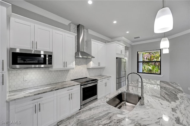 kitchen featuring white cabinets, wall chimney range hood, sink, appliances with stainless steel finishes, and decorative light fixtures