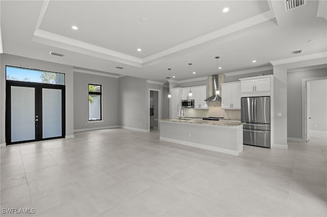 kitchen with wall chimney range hood, stainless steel appliances, an island with sink, white cabinets, and a raised ceiling