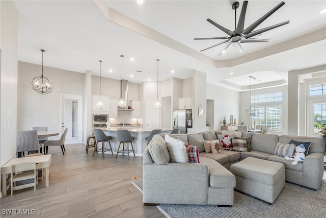 living room featuring ceiling fan with notable chandelier, a raised ceiling, light wood-type flooring, and sink