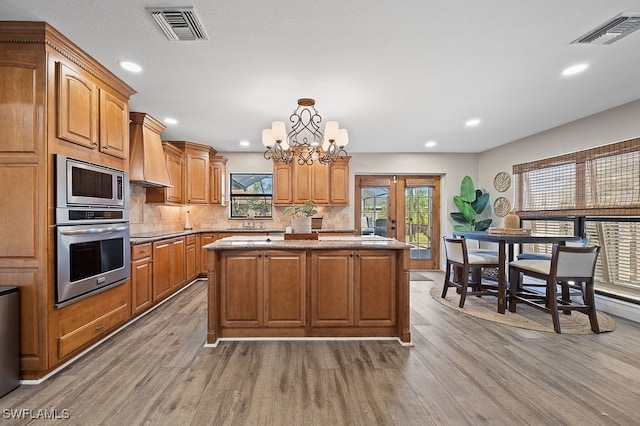 kitchen with stainless steel appliances, a notable chandelier, light hardwood / wood-style floors, a kitchen island, and custom range hood