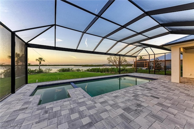 pool at dusk featuring a lawn, glass enclosure, a patio area, and a water view