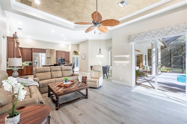 living room featuring visible vents, a high ceiling, a tray ceiling, light wood-style floors, and ceiling fan with notable chandelier