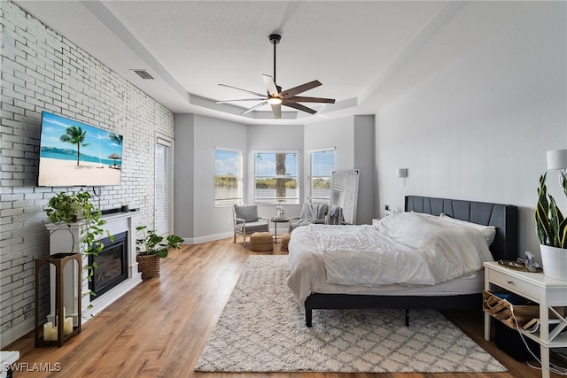 bedroom with light wood-style flooring, brick wall, visible vents, a raised ceiling, and a glass covered fireplace