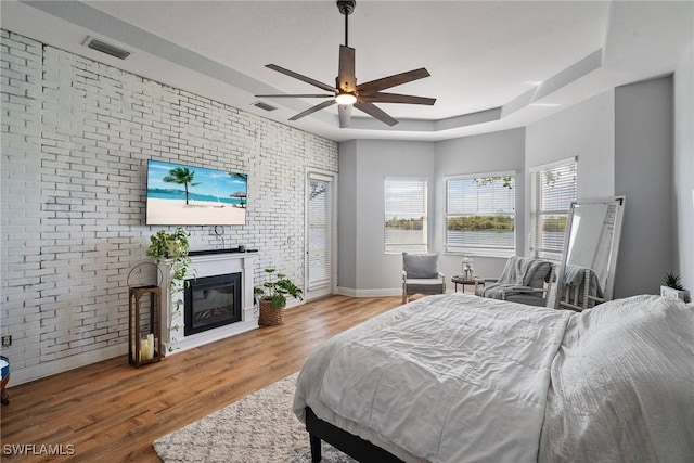 bedroom featuring a tray ceiling, visible vents, a glass covered fireplace, brick wall, and wood finished floors