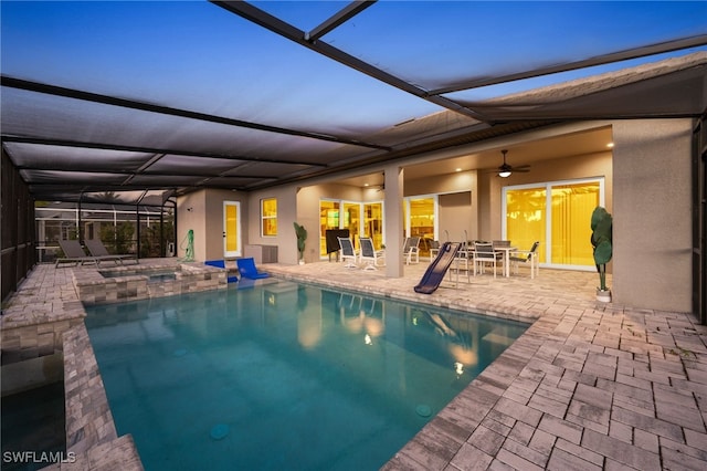 pool at dusk with a lanai, a patio area, ceiling fan, and a hot tub