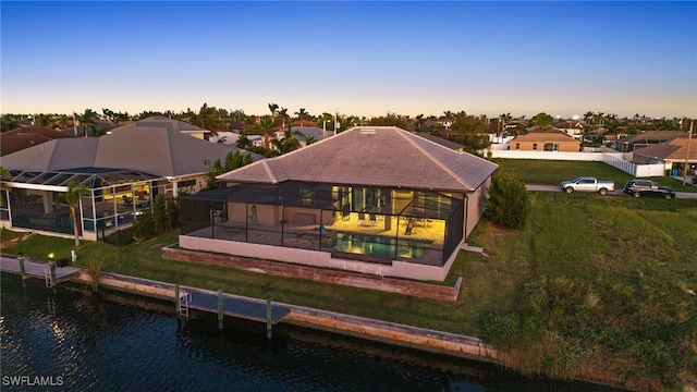 back house at dusk with glass enclosure, a water view, and a yard