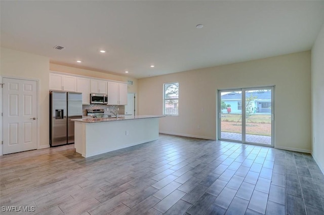 kitchen featuring light stone countertops, a kitchen island with sink, stainless steel appliances, decorative backsplash, and white cabinets