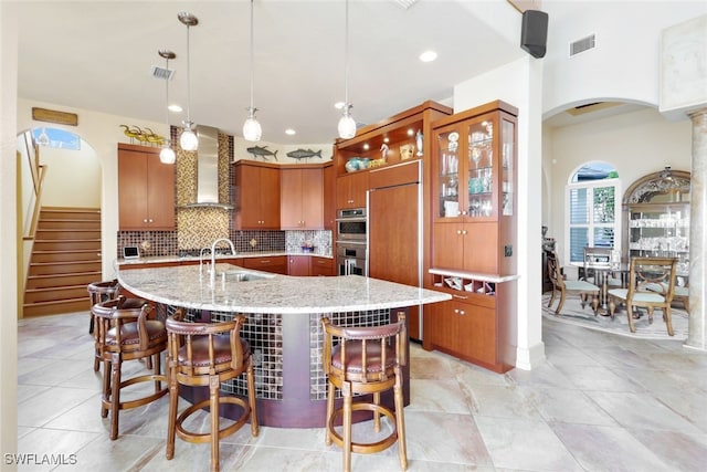 kitchen featuring wall chimney exhaust hood, paneled fridge, a spacious island, sink, and hanging light fixtures