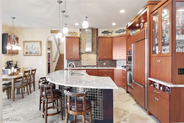 kitchen featuring sink, wall chimney exhaust hood, hanging light fixtures, and a center island with sink