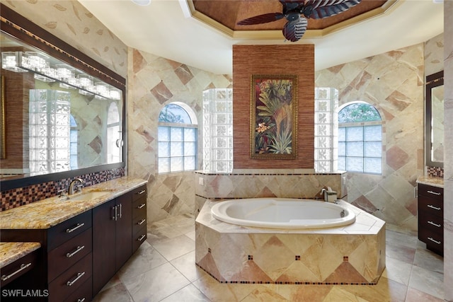 bathroom with vanity, a tray ceiling, ceiling fan, tiled tub, and tile walls