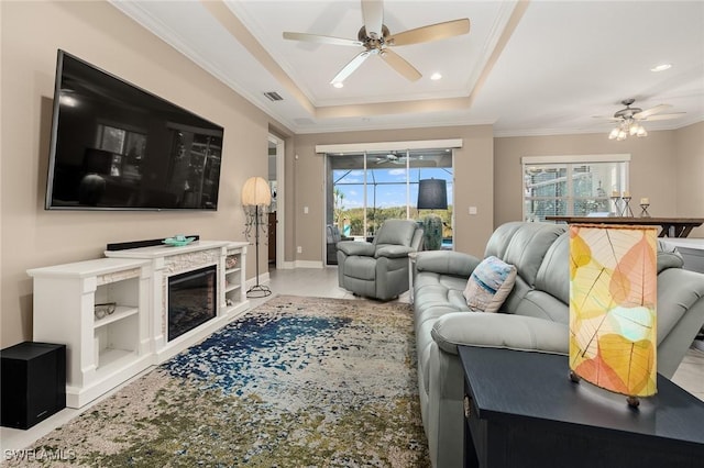 living room with a fireplace, a ceiling fan, visible vents, a tray ceiling, and crown molding