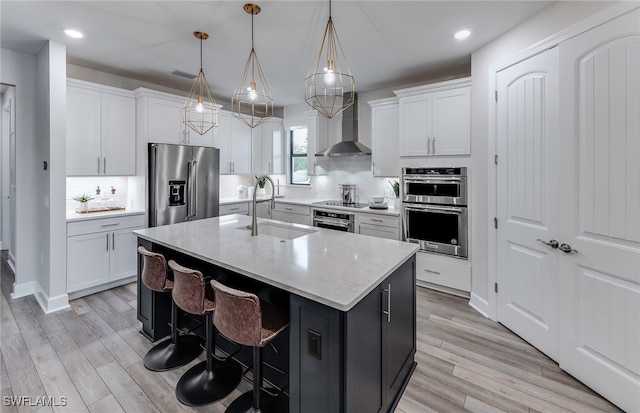 kitchen with sink, stainless steel appliances, wall chimney range hood, an island with sink, and decorative light fixtures