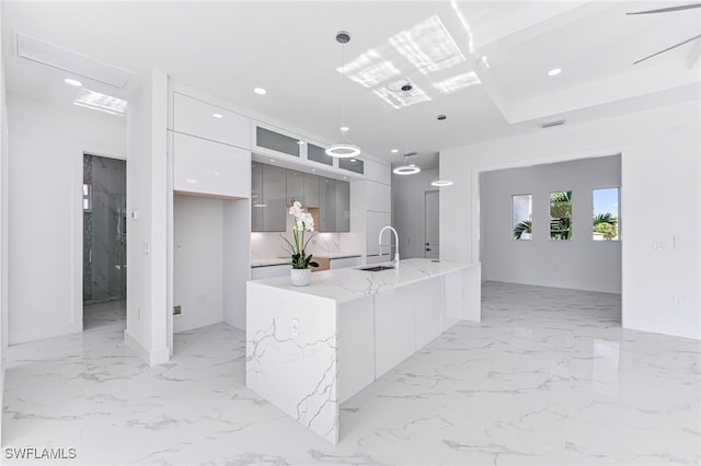 kitchen with a kitchen island with sink, white cabinets, sink, light stone countertops, and decorative light fixtures