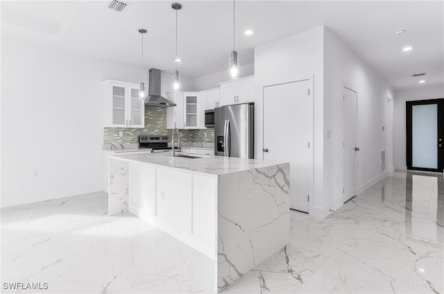 kitchen featuring appliances with stainless steel finishes, wall chimney range hood, a center island with sink, decorative light fixtures, and white cabinets