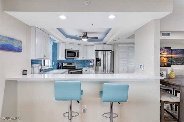 kitchen with kitchen peninsula, a tray ceiling, a kitchen bar, white cabinets, and appliances with stainless steel finishes