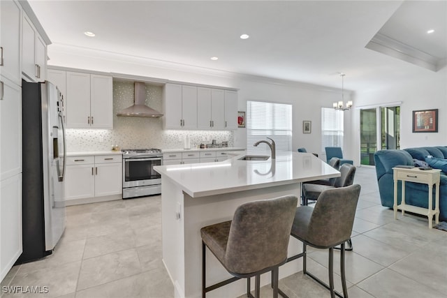 kitchen featuring appliances with stainless steel finishes, ornamental molding, wall chimney exhaust hood, decorative light fixtures, and a center island with sink