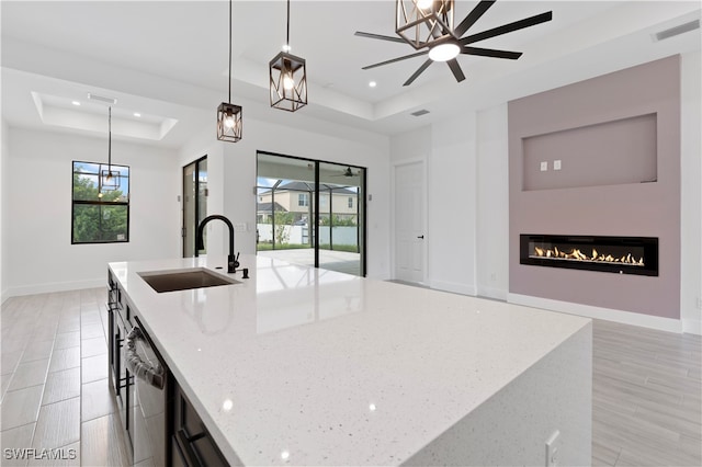 kitchen with light stone counters, a raised ceiling, sink, decorative light fixtures, and an island with sink