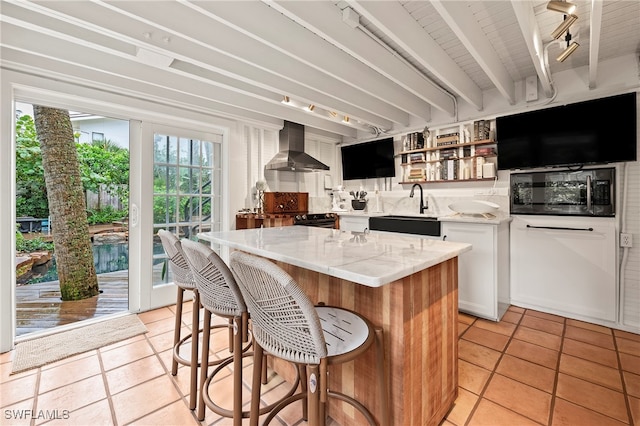 kitchen featuring light stone counters, wall chimney exhaust hood, sink, beamed ceiling, and a kitchen island