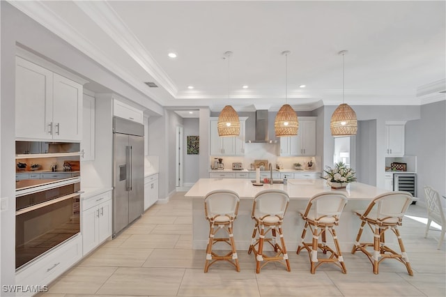 kitchen with stainless steel appliances, wall chimney range hood, pendant lighting, a center island with sink, and white cabinets