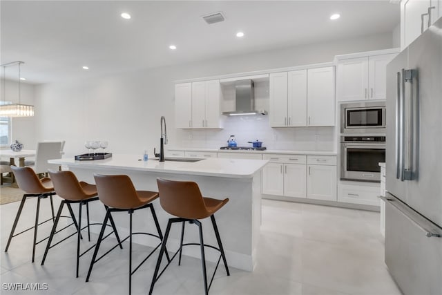 kitchen with wall chimney range hood, an island with sink, decorative light fixtures, a breakfast bar, and appliances with stainless steel finishes