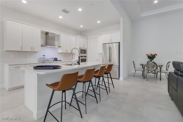 kitchen featuring white cabinets, appliances with stainless steel finishes, wall chimney exhaust hood, and an island with sink