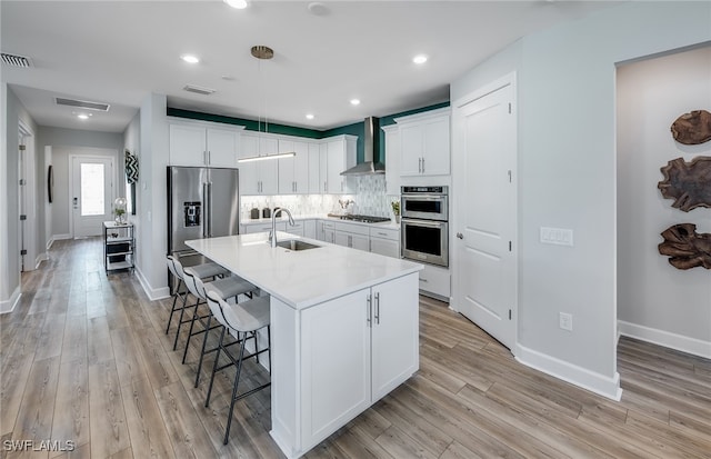 kitchen with sink, wall chimney exhaust hood, a kitchen island with sink, white cabinets, and appliances with stainless steel finishes