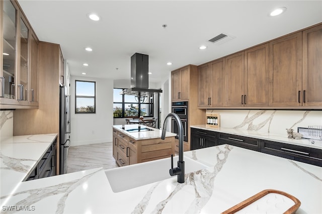 kitchen featuring light stone countertops, light hardwood / wood-style flooring, backsplash, island exhaust hood, and appliances with stainless steel finishes