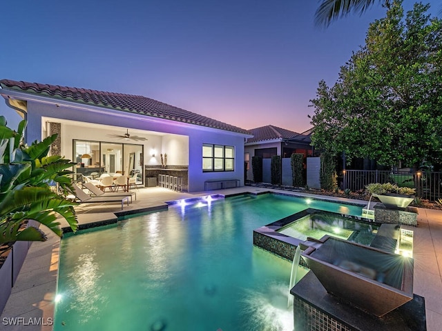 pool at dusk featuring a patio area, ceiling fan, pool water feature, area for grilling, and an in ground hot tub