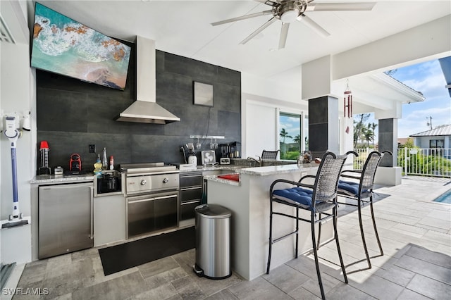 kitchen with light stone countertops, a kitchen breakfast bar, stainless steel refrigerator, and wall chimney exhaust hood