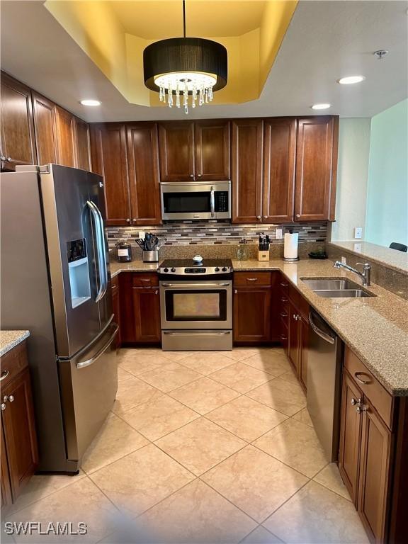 kitchen with a raised ceiling, sink, appliances with stainless steel finishes, decorative light fixtures, and light stone counters