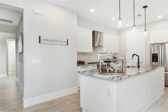 kitchen with wall chimney range hood, a center island with sink, light hardwood / wood-style flooring, stainless steel fridge with ice dispenser, and white cabinetry