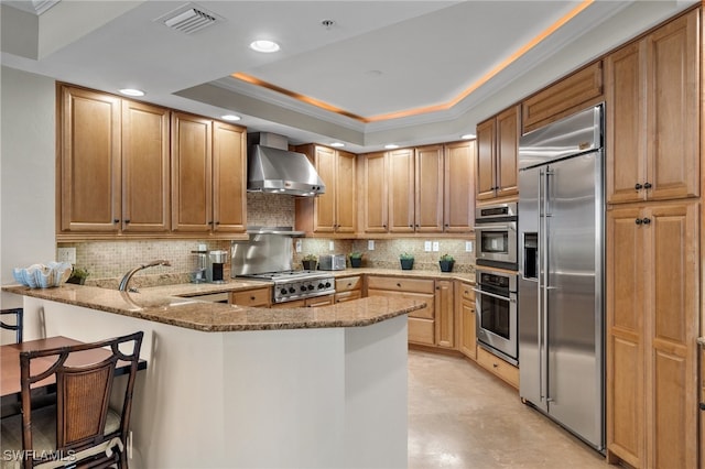 kitchen with kitchen peninsula, appliances with stainless steel finishes, light stone counters, a tray ceiling, and wall chimney range hood