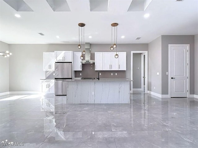 kitchen with wall chimney range hood, an island with sink, decorative light fixtures, white cabinetry, and stainless steel refrigerator