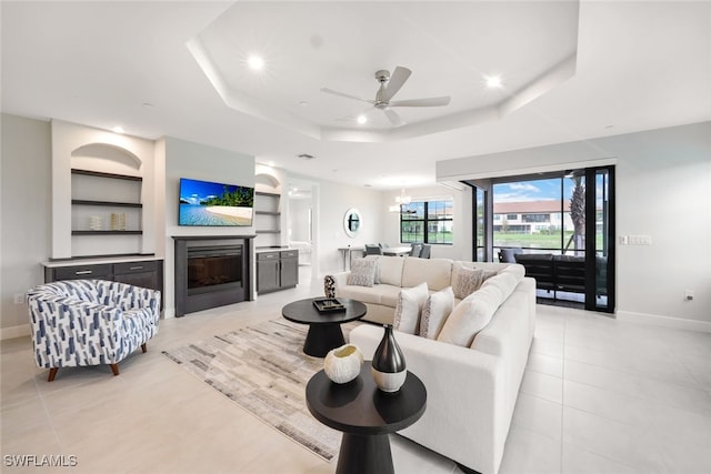 living room featuring a raised ceiling, built in shelves, light tile patterned floors, and ceiling fan with notable chandelier