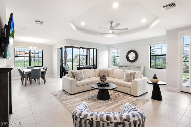 tiled living room with a tray ceiling, a wealth of natural light, and ceiling fan with notable chandelier