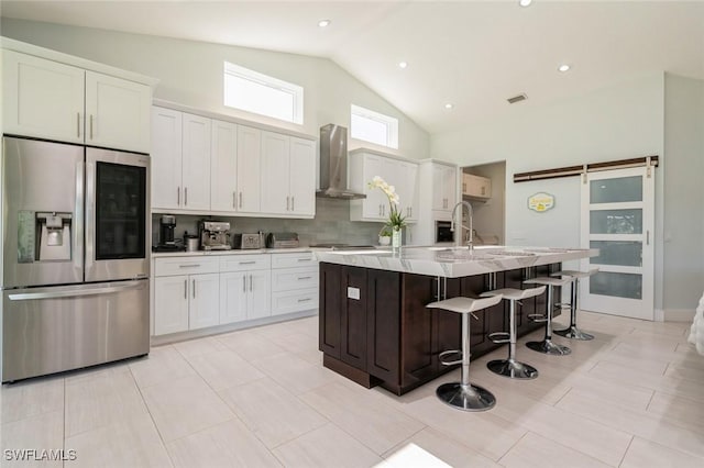 kitchen featuring stainless steel appliances, wall chimney range hood, a barn door, an island with sink, and white cabinets