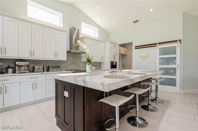 kitchen with white cabinets, wall chimney range hood, a barn door, an island with sink, and a kitchen bar