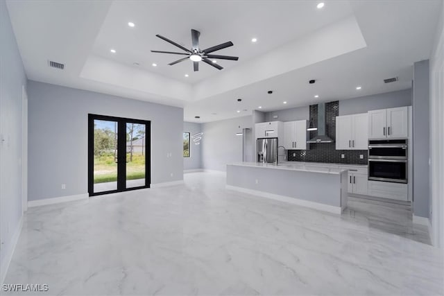 kitchen with a raised ceiling, wall chimney range hood, an island with sink, appliances with stainless steel finishes, and white cabinetry